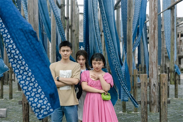 In A Year Without a Job, three characters stand among blue dyed cloths, a moment of their journey captured.