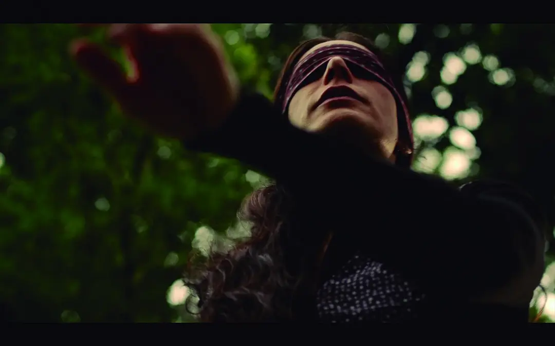 A woman blindfolded reaches out in a forest scene from "Através da Sombra".