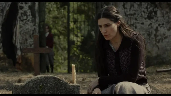 A woman sits somberly by a grave with a candle in "Através da Sombra".