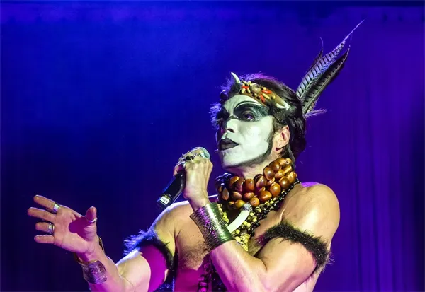 Costumed performer with face paint holds mic in "Homem com H".